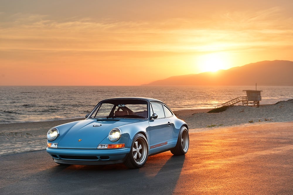 A blue classic Porsche parked beside the ocean at sunset